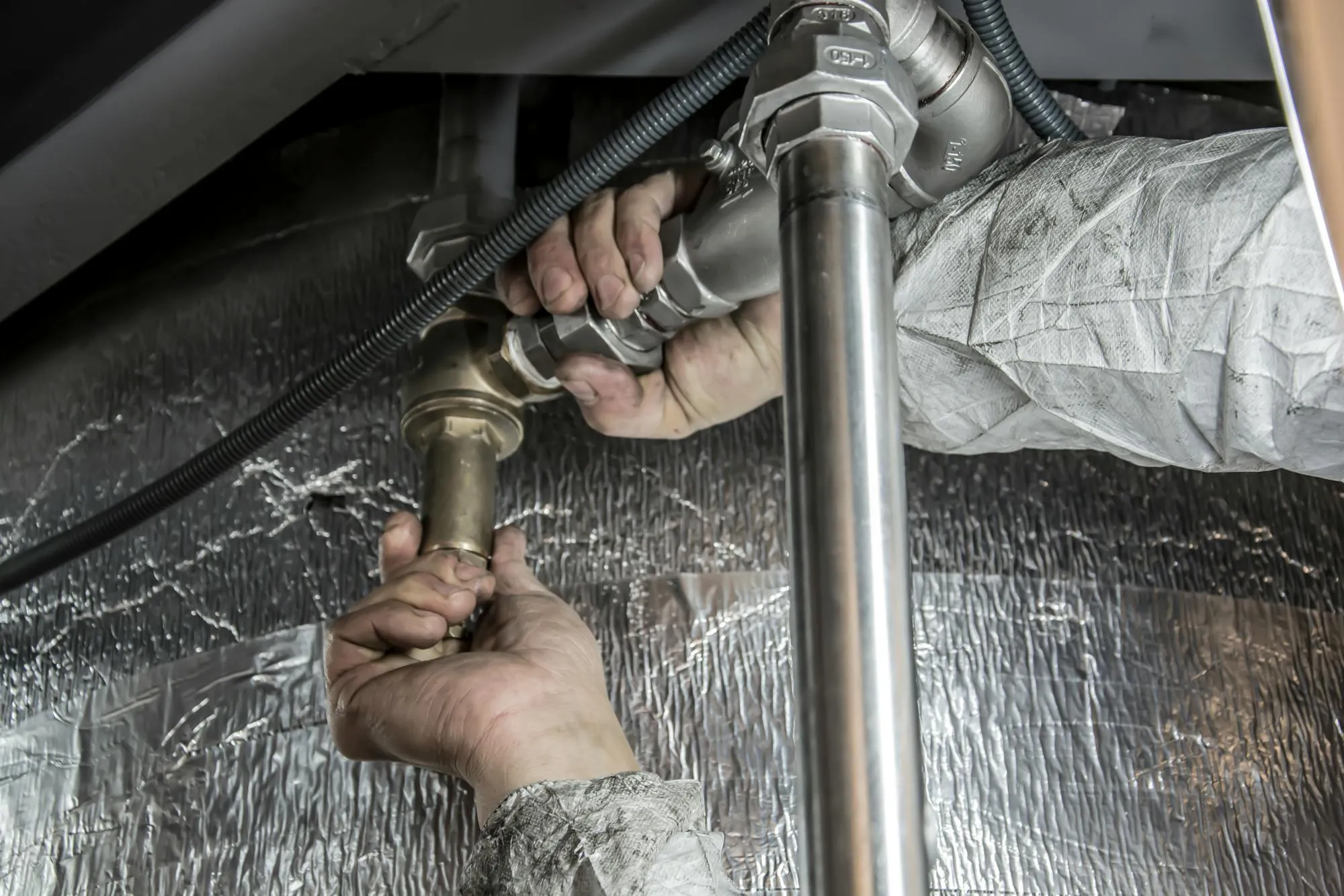 Hands fitting brass and copper pipework under a kitchen sink.